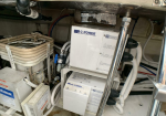 Compact boat engine room featuring a white C-POWER ISO TRANSFORMER panel, bundled hoses, and metal piping along a concrete floor.