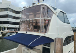 White motor yacht docked at a marina with a blue aft sunshade extended over the deck and a transparent aft cockpit enclosure; modern building in the background.