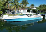 A white and blue motor yacht docked in a calm canal beside a tropical backyard with palm trees and a white house behind it.