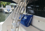 Stainless steel folding ladder attached to the back of a white boat, with a blue duffel bag resting on the bench nearby. A marina with other boats is visible in the background.