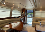 Interior of a luxury yacht cabin with a long beige sofa, wooden table, and cream blinds lining the windows.