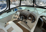 Boat helm with steering wheel and gauges inside a marina cockpit, view of yachts outside.