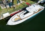 Bird's-eye view of a white luxury yacht docked along a canal, with palm trees, a red wooden walkway, and a pool area nearby