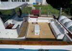 Open yacht deck with white seating, beige teak flooring, and wooden trim; boat moored near a grassy yard in background.