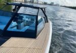 Sleek boat with a glass-cabin cockpit and wooden deck gliding on a rippling river with a city skyline in the distance.
