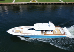 White luxury motor yacht cruising near a grassy shoreline, creating a wake in blue water with a spacious teak deck and seating area at the stern.