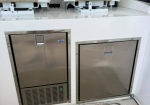 Two stainless steel marine appliances beneath seats: a fridge with a vented lower panel on the left and a separate freezer on the right.