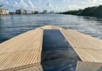Perspective from the bow of a wooden decked boat looking toward a city skyline across calm water.