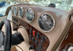 Close-up of a yacht helm with round analog gauges set in a wood-paneled dashboard, steering wheel to the left, marina visible through the windshield.