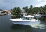 A white motor yacht cruises through a calm waterway with palm trees and waterfront houses in the background.
