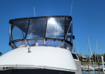 Close-up of a white yacht's flybridge and windshield at a marina, with a clear blue sky and sailboat masts in the background.