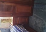 Wooden bedroom wardrobe and nightstand beside a bed with blue-gray striped bedding; a gold textured lamp sits on the nightstand.