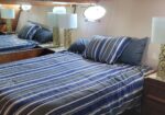 Boat cabin bedroom with a striped blue-gray bed, matching pillows, and two gold-patterned lamps on nightstands beside the bed