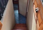 Narrow boat stairwell with beige walls, wooden cabinets, and a left-side control panel of switches, leading to a doorway at the bottom.