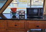 Boat galley with microwave and toaster on a black granite counter, orange and teal bowls, and a small potted bonsai, overlooking a marina.