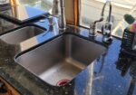 Double stainless steel kitchen sink on a black granite counter with a soap dispenser, sponges, and scrub brush nearby by the window.