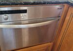 Built-in stainless steel Miele dishwasher under a dark granite countertop, surrounded by wooden cabinets.