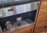 Built-in stainless steel Miele coffee machine in a wooden kitchen cabinet with a metal milk pitcher on the drip tray