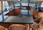Boat kitchen/gally interior with dark granite counters, wooden cabinetry, and a central island, overlooking the water through large forward windows.