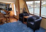 Interior of a luxury yacht: wooden cabinetry, curved glass bar, and wicker stools, with large windows showing the marina outside.