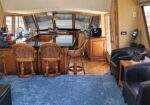 Boat interior: cafe-style galley with granite island, wooden cabinets, and wicker bar stools facing large windows looking out at a marina.